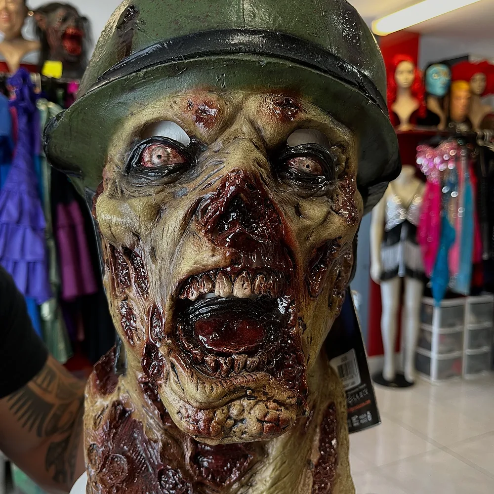 Zombie soldier latex mask with a battered helmet, held up in the shop with wigs and costumes behind