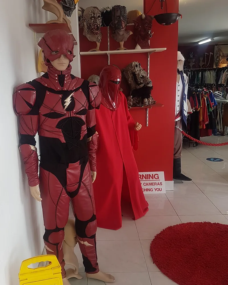 Red armoured superhero suit on a mannequin beside a red hooded guard robe, with horror masks on the shelves behind